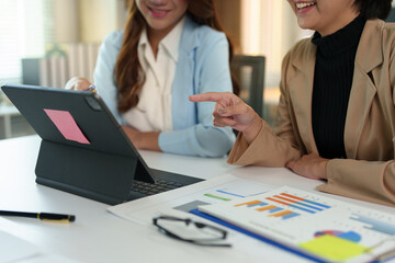Two young businesswomen pointing to plans, statistics, and data graphs and explaining the conditions of their partnership, happily acknowledging their turnover and income during negotiations.