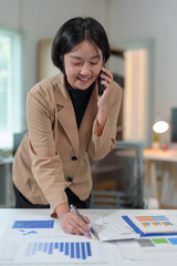 Attractive Asian businesswoman standing on the phone talking about real estate projects and chatting with happy smiles at the office.