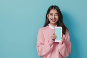 Fototapeta premium Full body young smiling happy woman of Asian ethnicity 20s in pink sweater stand near big mobile cell phone with blank screen workspace area chatting isolated on pastel plain light blue background