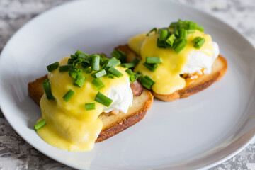 Delicious English breakfast. Eggs benedict with sauce and bacon on toasted white bread on a gray plate, close-up on a light background