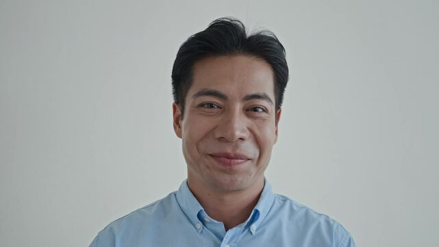 Close up portrait of cheerful Asian guy in blue casual shirt nodding and smiling at camera on white background