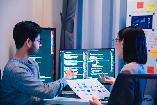 Asian software developers teams working on multiple screens displaying code and application diagrams at night in modern office