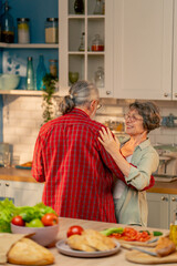 close up in bright kitchen an elderly woman in a light green shirt with her husband in an apron standing in the kitchen hugging smiling romantically dancing