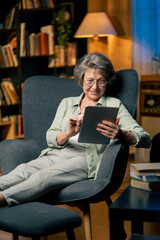 warm light living room with library big blue armchair elderly woman sitting watching a news from tablet modern grandma