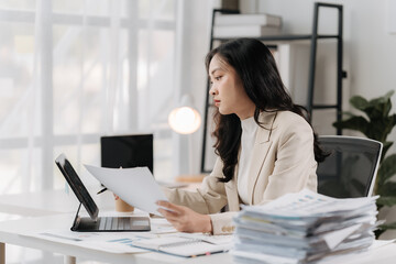 Fototapeta premium Focused Determination: A young businesswoman meticulously examines paperwork in her modern office, her expression reflecting dedication and focus amidst a stack of documents.
