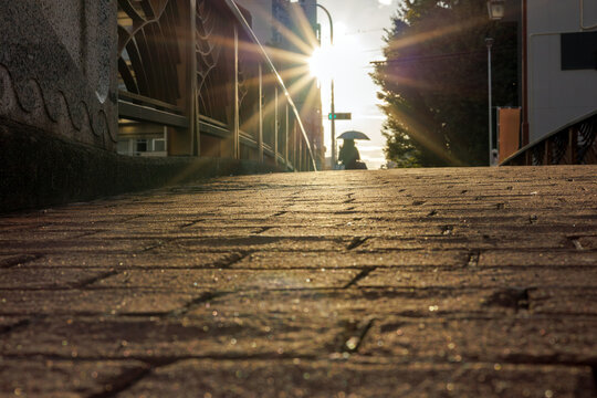 The silhouette of a person with a sun umbrella is seen on the far side of a cobblestone paved pedestrian bridge. The scene creates a sense of summer heat with long afternoon shadows and a sun flare.