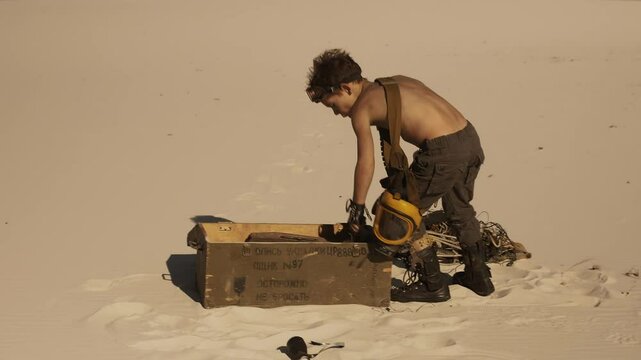 A young boy dressed as a post-apocalyptic warrior scavenges through a box of junk in a desert wasteland. Ideal for survival, dystopian themes, adventure stories, and creative media projects. High