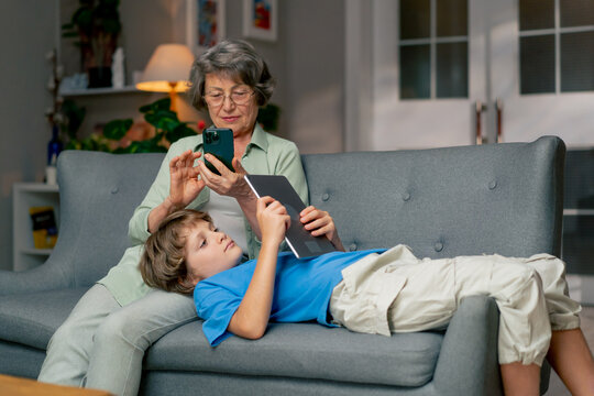bright living room with sofa grandmother sitting on sofa grandson lying and watching cartoons on tablet happy family - Powered by Adobe