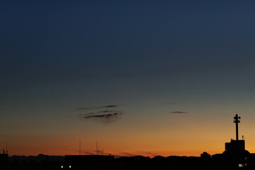 Fototapeta premium In a gradient dusk sky full of orange and blue hues, Venus shines faintly high above the silhouettes of buildings and antennas.