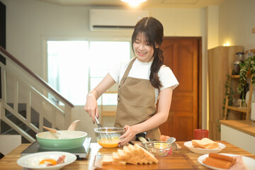 Young asian woman wearing an apron whisking eggs in a glass bowl at kitchen counter