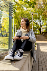 beautiful woman in wireless headphones with a phone on the street in autumn, using smartphone and headphones,  listen  to music, sunny autumn day 