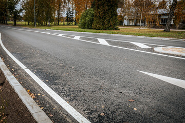 Obraz premium A quiet, empty asphalt road with a white directional arrow and painted lane markings. The scene shows fall foliage and trees in the background, with a nearby building partially visible.