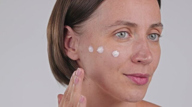 Skincare ads. Portrait female face. Caucasian woman smile apply Moisturizer Sunscreen SPF, putting a base foundation for makeup. Daily morning beauty routine. Isolated Studio Light Grey background. 