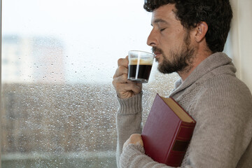 Young adult man enjoys hugging a book and having a cup of warm coffee in his living room at home next to the window on a rainy day, warm at home in autumn or winter, enjoying reading, reading book