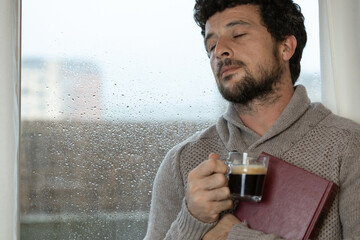 Young adult man enjoys hugging a book and having a cup of warm coffee in his living room at home next to the window on a rainy day, warm at home in autumn or winter, enjoying reading, reading book