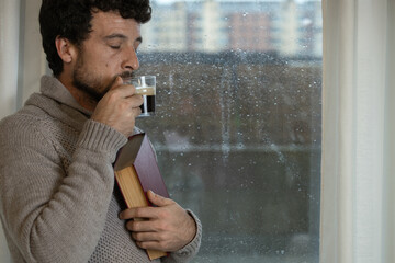 Young adult man enjoys hugging a book and having a cup of warm coffee in his living room at home next to the window on a rainy day, warm at home in autumn or winter, enjoying reading, reading book