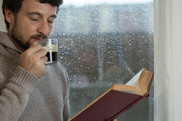 Young adult man enjoys reading a book and having a cup of warm coffee in his living room at home next to the window on a rainy day, warm at home in autumn or winter, enjoying reading, curtains