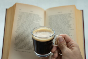 Young adult man holds a cup of warm coffee with his hand along with a book, warm at home in autumn or winter, close-up cup of coffee and novel book background
