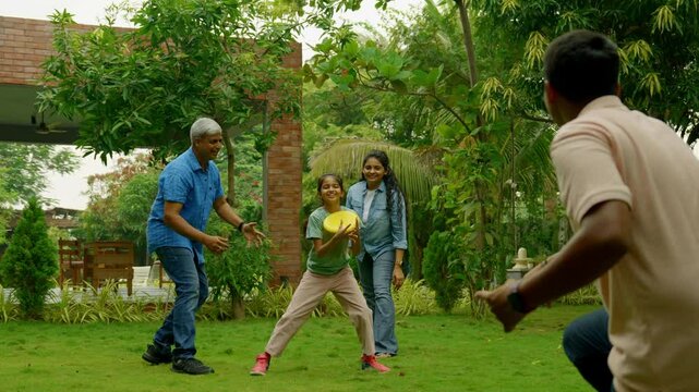 Indian Asian family of four spending quality time outdoors in their home lawn, parents in their forties watching as teenage son teaches his little sister how to throw a frisbee or flying disc