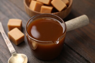 Tasty caramel sauce in glass measuring cup, sweet candies and spoon on wooden table, closeup