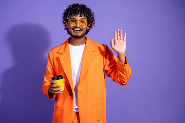 Photo of handsome good mood guy wear orange suit enjoying tea waving hi emtpy space isolated violet color background © deagreez