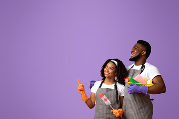 Cheerful Black Couple Holding Cleaning Supplies And Pointing At Copy Space Over Pink Background, Free Place For Your Ad