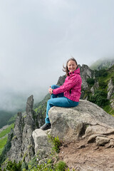 Happy smiling hiker in pink jacket and blue pants sits on large rock at edge of cliff. Behind her, rugged rock formations and green slopes are partially veiled in mist.