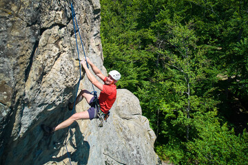Obraz premium Aerial view of male rock climber ascending rugged limestone cliff with harness and rope for safety. Sportsman climbing on vertical large boulder at Dobvush Rocks in Carpathian mountains, Ukraine.