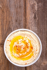 A bowl of creamy hummus drizzled with olive oil and sprinkled with paprika, over rustic wooden background. Space for text.