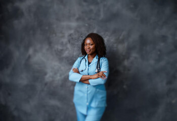 A woman with braided hair stands with her arms crossed in front of a gray wall, wearing a blue medical uniform and a stethoscope.