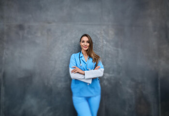 A woman in a blue medical scrubs stands against a textured gray wall with her arms crossed. She is smiling and looking directly at the camera.