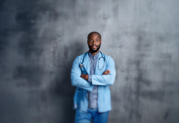 A man in a blue lab coat with a stethoscope around his neck stands against a gray textured wall with his arms crossed.