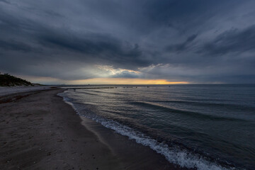 a peaceful evening at the beach in Warnemünde, Rostock, Germany, with gentle waves lapping against the shore under a moody sky, as the sun sets in the distance over the Baltic Sea