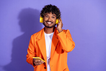 Photo of handsome good mood guy wear orange suit listen music earphones modern gadget emtpy space isolated violet color background