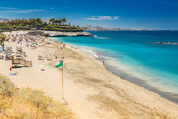 Fototapeta premium A sunny scene at Playa del Duque, Tenerife, with swimmers and sunbathers fill the beach, with a green flag signaling safe swimming