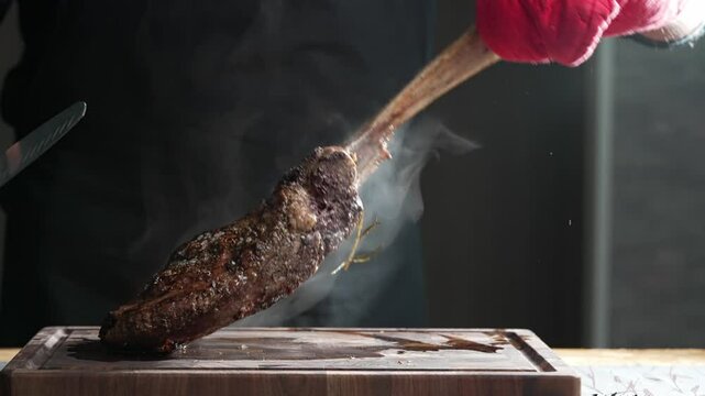 Chef wearing black gloves is holding a smoking tomahawk steak on a wooden board
