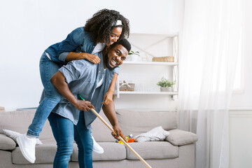 Joyful Spring-Cleaning. Playful African American Couple Fooling Together During Tidying Flat, Woman Piggybacking Her Boyfriend While He Mopping Floor