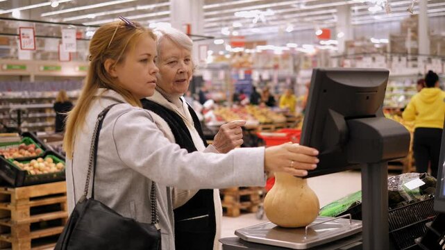 A social worker helps an elderly woman weigh a pumpkin in a large supermarket. A woman and her grandmother weigh a pumpkin on an electronic scale in a grocery store.