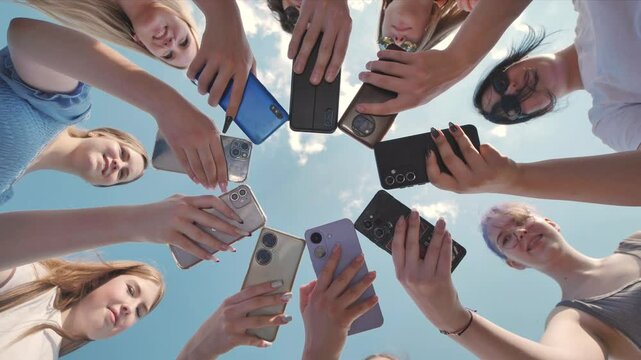 Group of teenage girls and boys holding smartphones making a circle shape with their mobile phones against the blue sky.