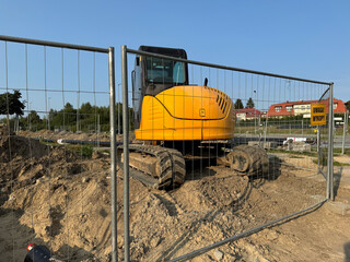 A yellow excavator parked behind a metal fence on a construction site with warning signs