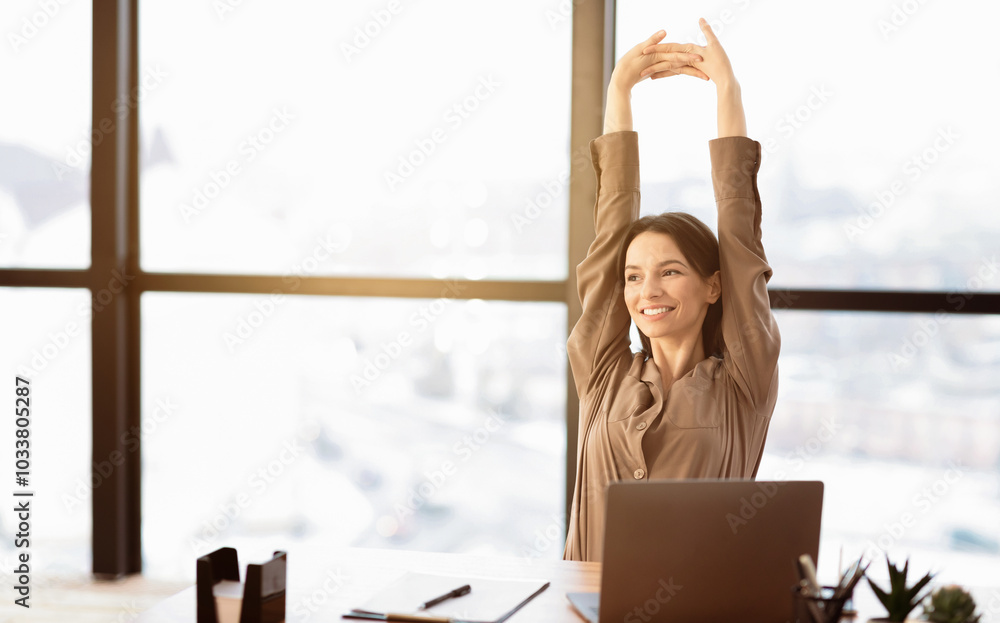 Wall mural smiling young woman in brown long sleeve shirt stretching in front of desk at office, looking aside,