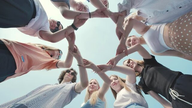 Group of young students forming a circle with their hands together outdoors, low angle view.