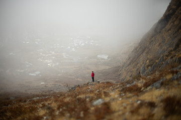 A person in a red jacket stands on a misty hillside overlooking a foggy valley.