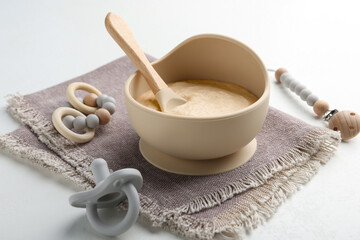 Delicious baby food in bowl with spoon, pacifier and teether on white background