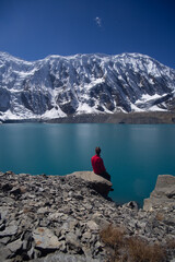 A person in a red jacket sits on a rock, gazing at the serene blue lake and snow-capped mountains...