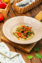 Dish of green beans with tomatoes and bell peppers in white sauce, in a plate, cafe, restaurant, on a wooden table.