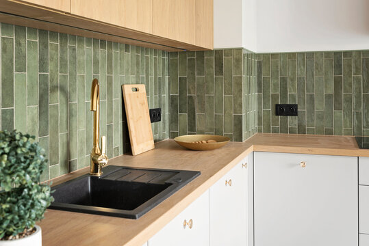 Kitchen interior in modern style with kitchen sink with tap, contemporary green tile on the wall, wooden counter and furniture. New apartment with design decor. Real estate.
