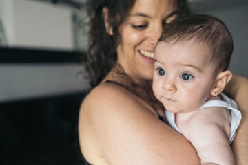 Young mother holding her baby girl at home. Young mother smiling while holding her baby girl at home looking through the window.