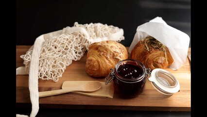 Flaky croissant next to jar of jam, woven bag, resting on wooden surface. Soft lighting enhances texture of pastry, evoking cozy morning meal with homemade, artisanal touch.