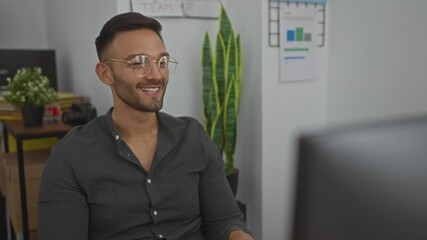 Young man, hispanic, smiling, wearing glasses in an office workspace, making a thumbs-up gesture while on a video call, indoor, modern, technology, plants, desk in background, charts, casual attire